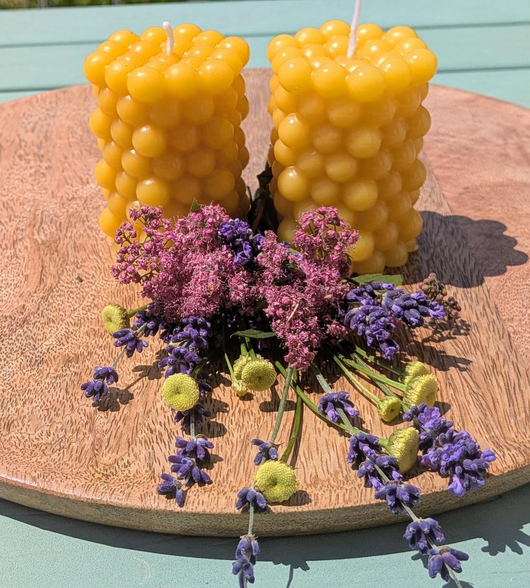 Zwei Bienenwachskerzen in Hexaeder-Form mit Kugelnoppen auf Holzscheibe mit Trockenblumen