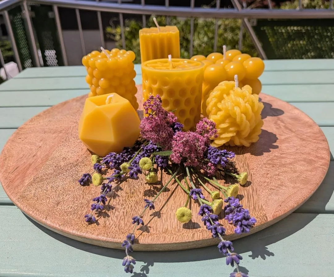 Sechs Bienenwachskerzen in verschiedenen Formen auf einem Holzteller, davor Lavendel