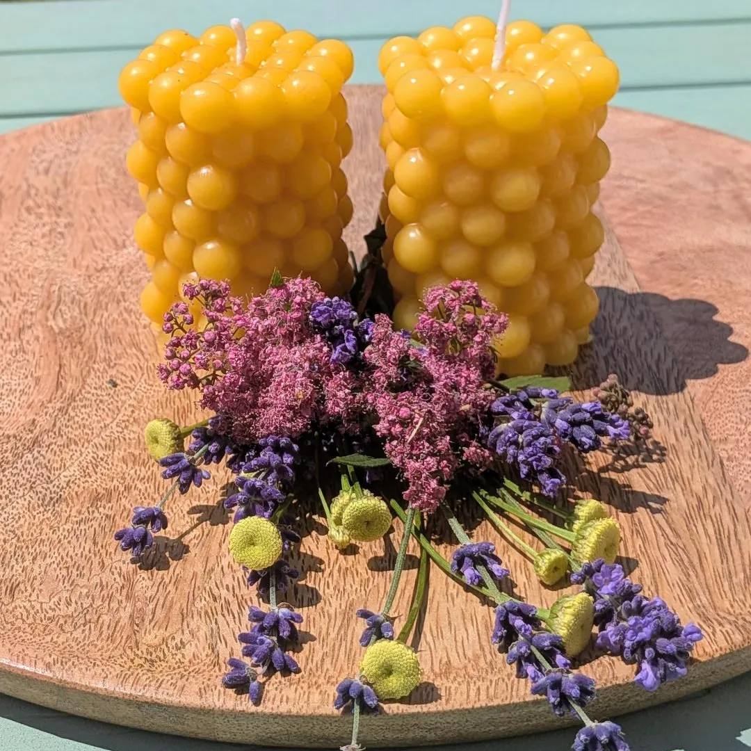Zwei Bienenwachskerzen mit Kugelnoppen-Struktur auf Holzscheibe, mit Lavendel und rosa Blüten