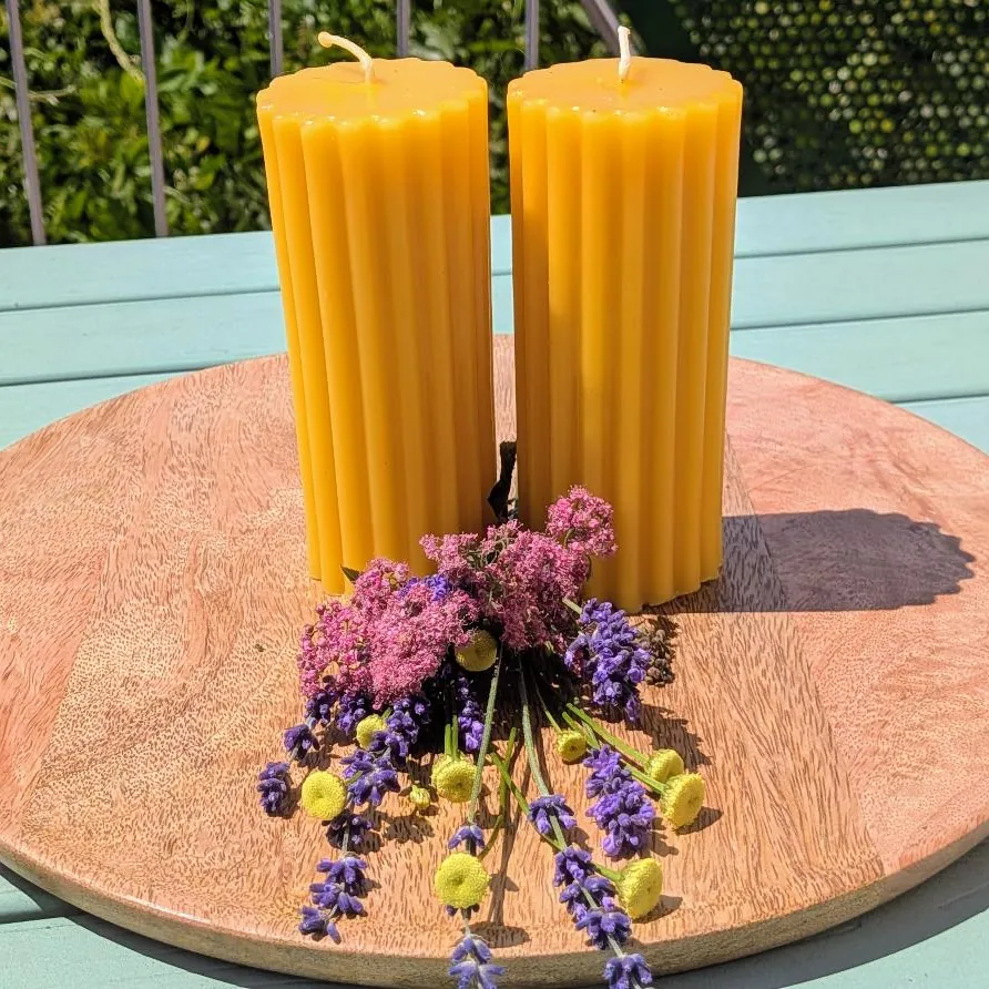 Zwei große Bienenwachsstumpen mit breiten runden Längsrillen auf Holzscheibe, mit Lavendel und rosa Blüten