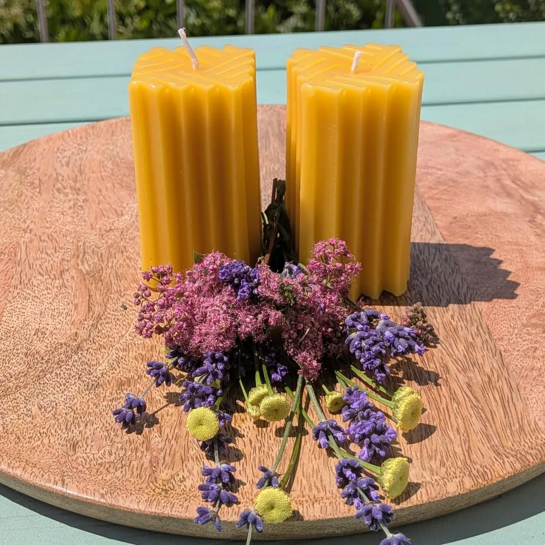 Zwei Bienenwachsstumpen mit senkrechten Rillen und gestuftem, eckigem Kopf auf Holzscheibe, mit Lavendel und rosa Blüten