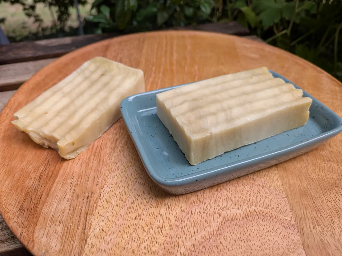 Zwei cremefarbene, geriffelte Stücke Arganöl-Olivenölseife auf Holzbrett, eines in einer blauen Seifenschale