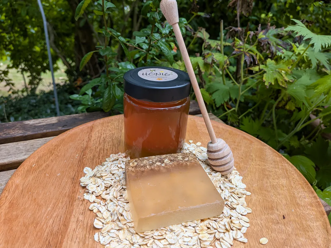 Honig- und Haferflockenseife auf einem Bett aus Haferflocken, dahinter ein Glas Sommertrachthonig mit Honiglöffel