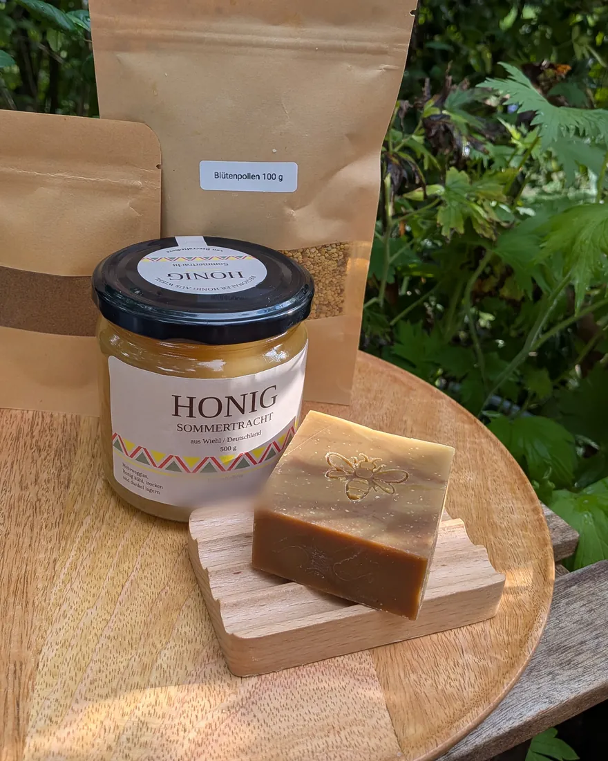 Honigseife mit Bienenprägung auf einer Holzschale, dahinter ein Glas Sommertrachthonig und ein Beutel Blütenpollen