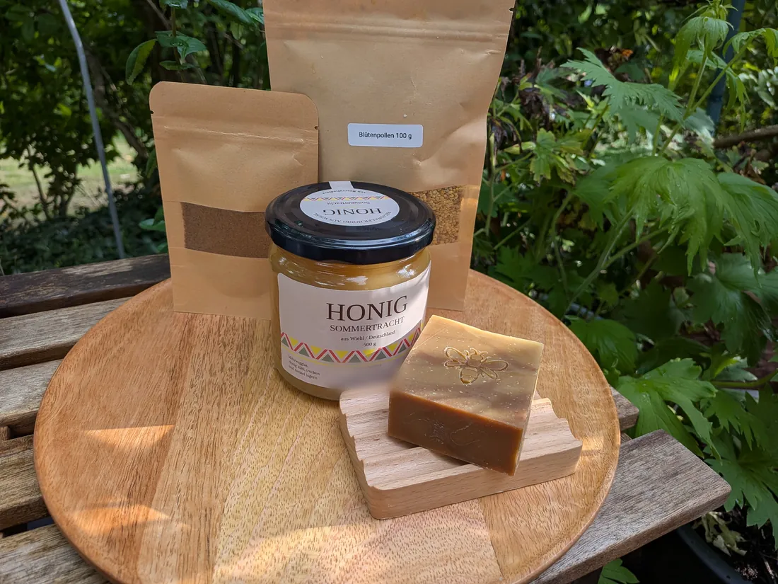 Honigseife mit Bienenprägung auf Holzschale, dahinter ein Glas Honig, ein Beutel Blütenpollen und ein Beutel Propolis