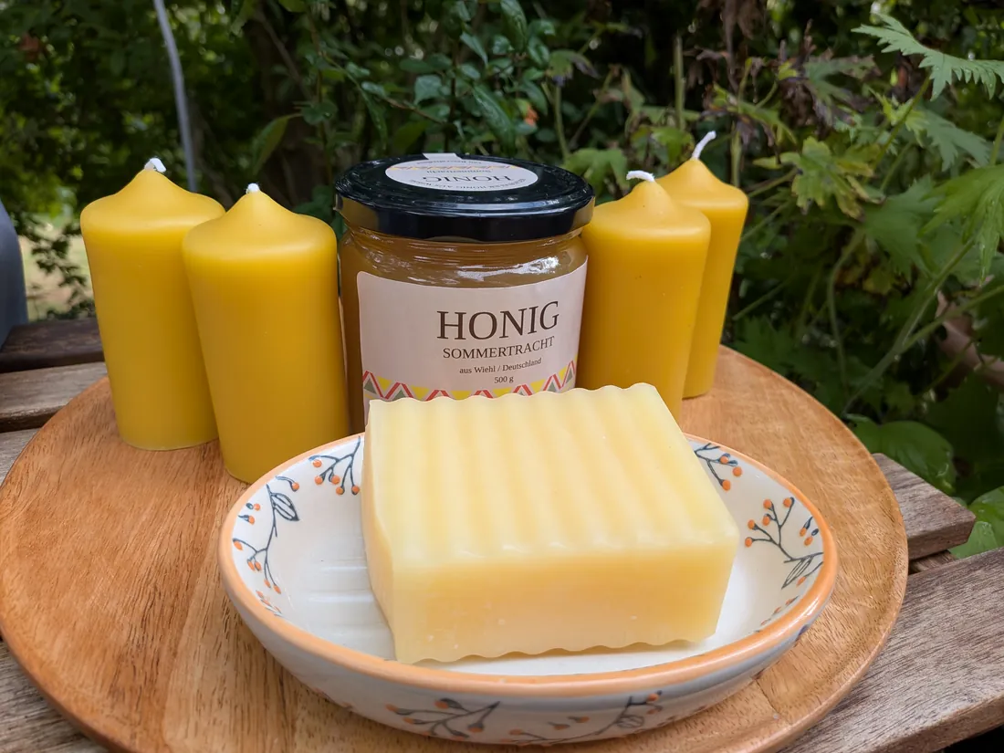 Vier Bienenwachskerzen, ein Glas Sommertrachthonig und eine Seife auf einer Schale, auf einem Holzbrett im Garten