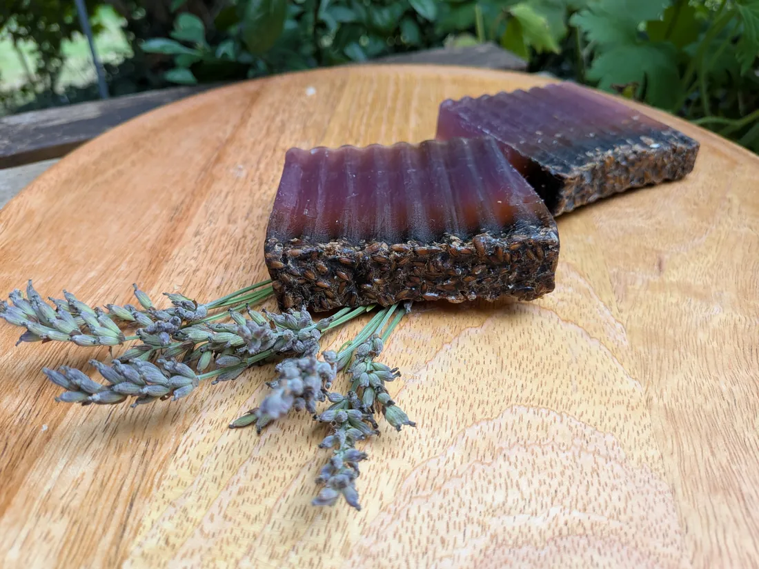 Zwei dunkle Stücke Lavendelseife mit geriffelter Oberseite und Lavendelblüten-Schicht auf Holzbrett, daneben Lavendelrispen