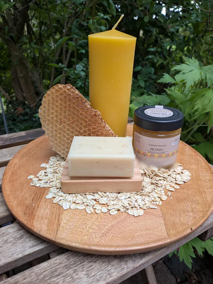 Große Bienenwachs-Stumpenkerze, Wabenstück, Honigglas und Honig-Haferflockenseife auf Holzschale zwischen Haferflocken