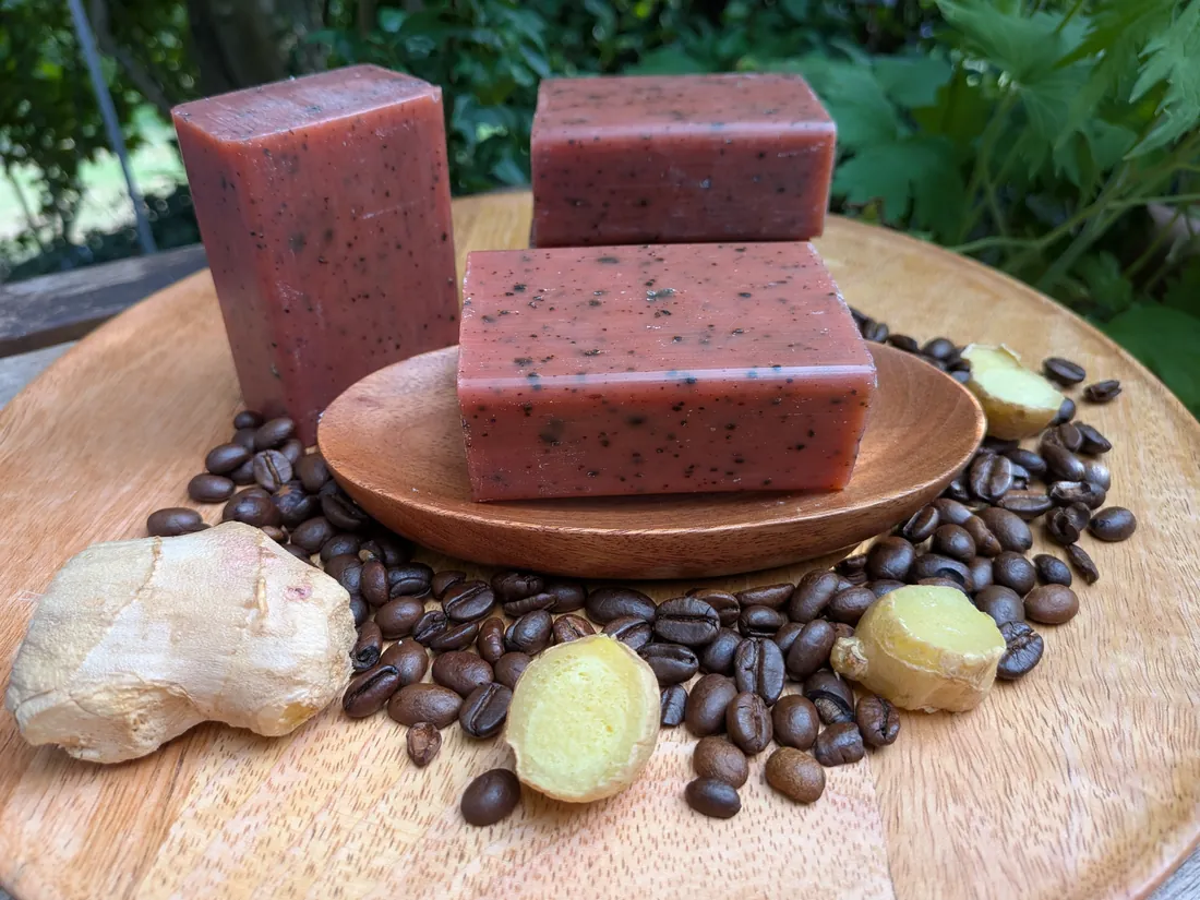 Drei Stücke rötliche Tonseife mit dunklen Kaffeepartikeln, eines auf einer Holzschale, daneben Kaffeebohnen und Ingwer