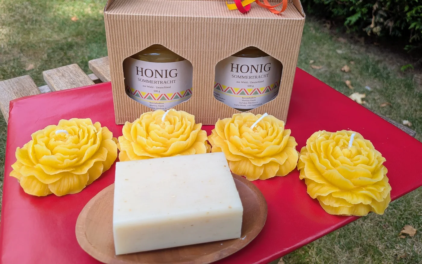 Geschenkkarton mit zwei Gläsern Sommertrachthonig, davor vier Rosenkerzen aus Bienenwachs und eine Honigseife auf einer Holzschale